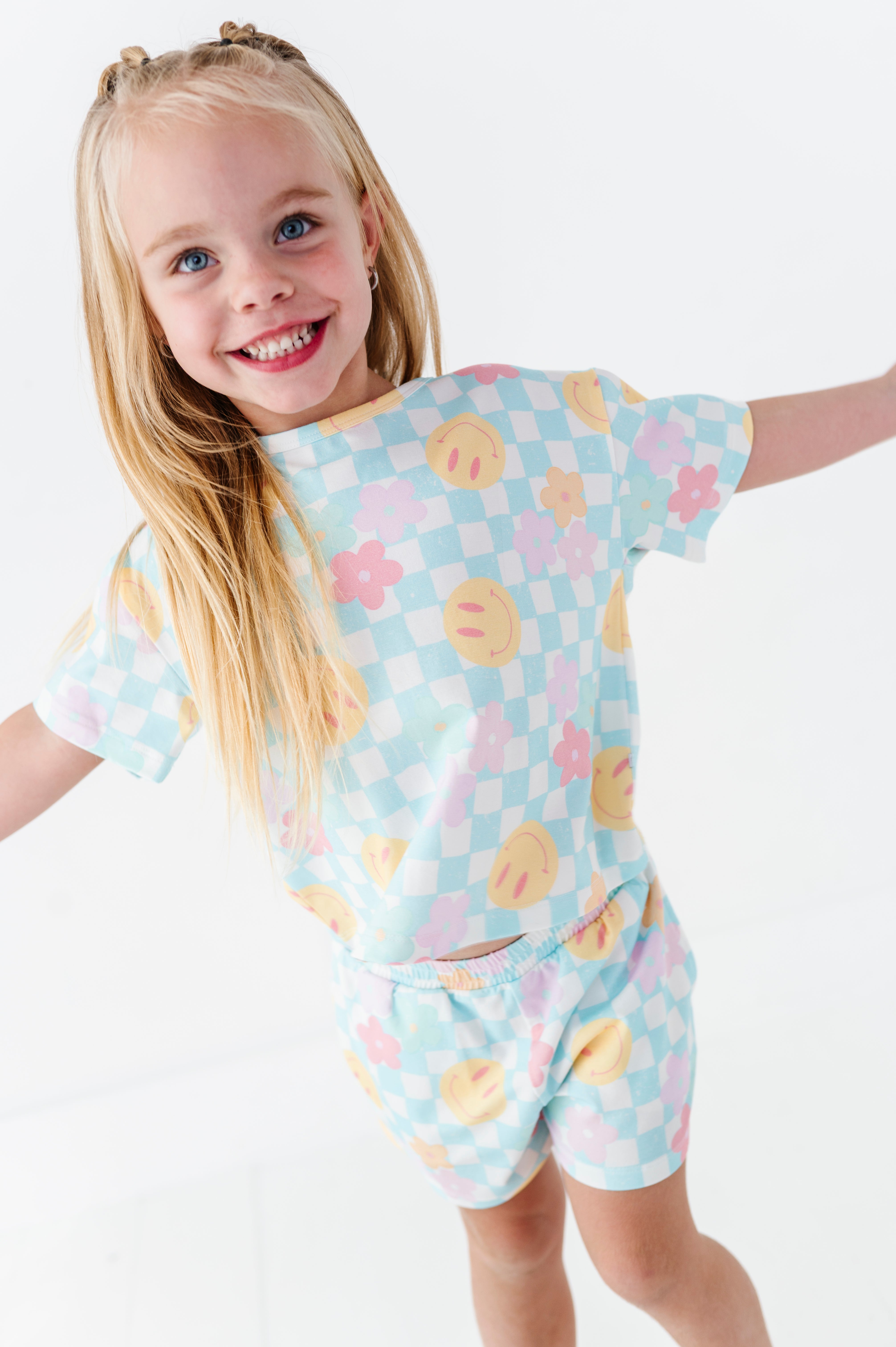 A young girl with blonde hair smiles while wearing a light blue checkered pajama set with yellow smiley faces and pink flowers, posed against a clean white background.
